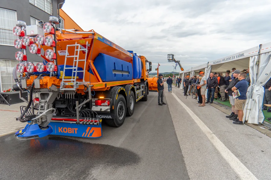 Oranžovo-modrý sypač na podvozku Scania projíždí kolem diváků u stanu s nápisem „TECHNIKA PŘIJEDE ZA VÁMI.“