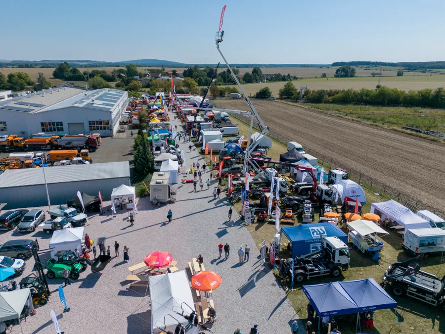 Štěrková ulička mezi stánky, návštěvníci v pohybu a vysoko vysunutá montážní plošina nad expozicí