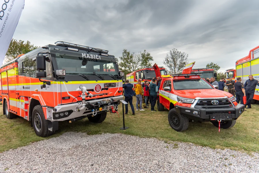 Hasičská cisterna na terénním podvozku a červený pickup Toyota s majákem stojí na travnaté ploše