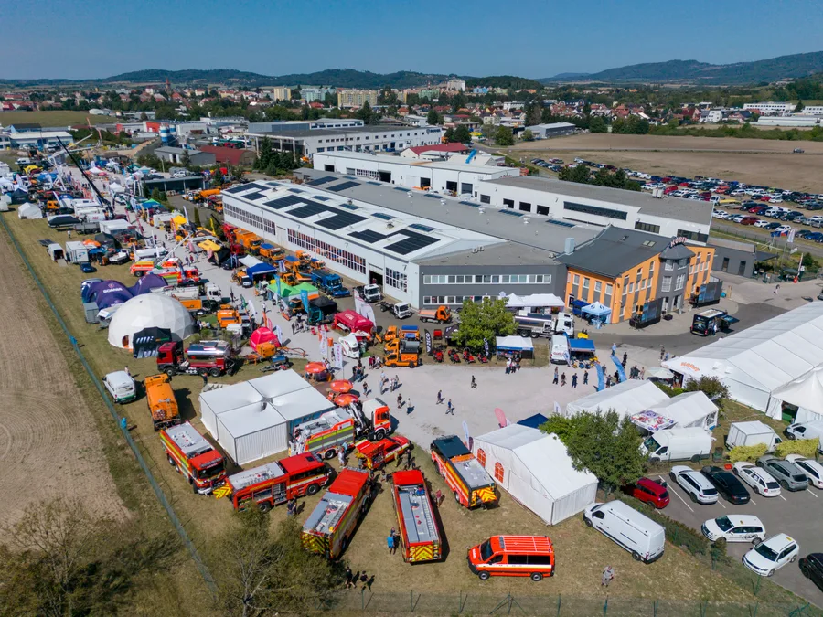 Hasičská technika v popředí, za ní hala s fotovoltaikou, stany a aleje strojů, v pozadí Jičín