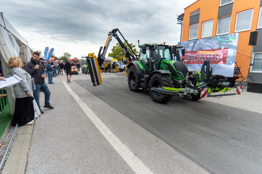 Zelený traktor s vyloženým výložníkovým mulčovačem projíždí kolem moderátora u diváckého stanu