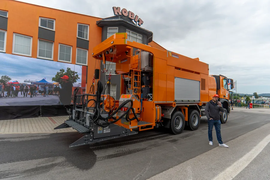 Oranžová silničářská nástavba se sklopenou zadní plošinou a hydraulikou před budovou KOBIT