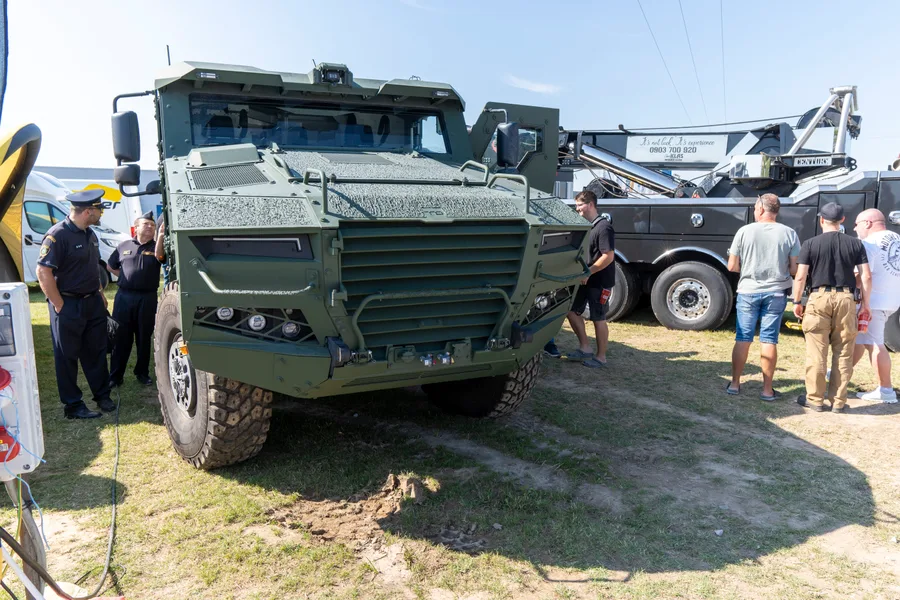 Zelené obrněné terénní vozidlo na trávě, vlevo dva muži v uniformách, vpravo skupina návštěvníků