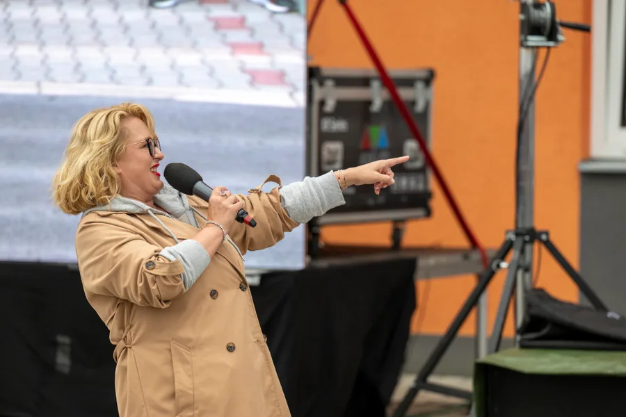 Moderátorka v béžovém kabátu drží mikrofon a ukazuje rukou do strany před LED obrazovkou