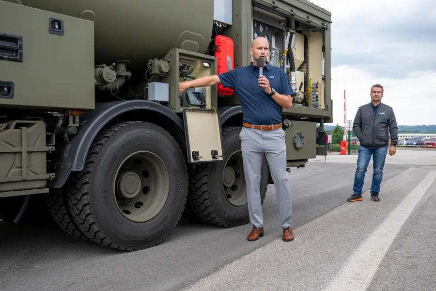 Muž s mikrofonem ukazuje na otevřenou technologickou skříň s hadicemi a manometry na boku vojenské cisterny