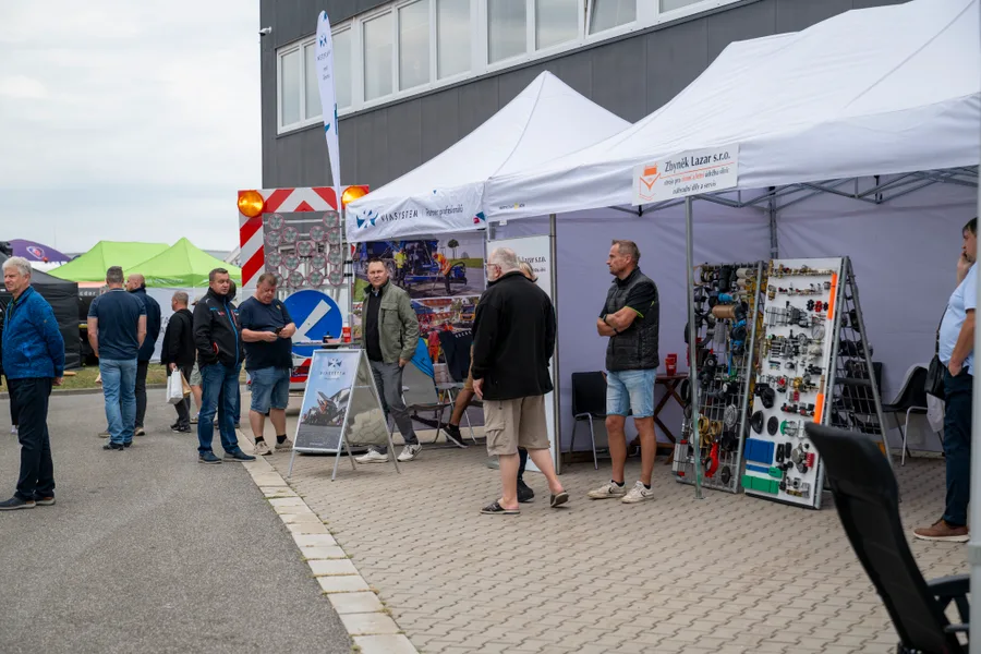 Návštěvníci u řady bílých stánků podél haly, vpravo panel s hydraulickými a strojními díly