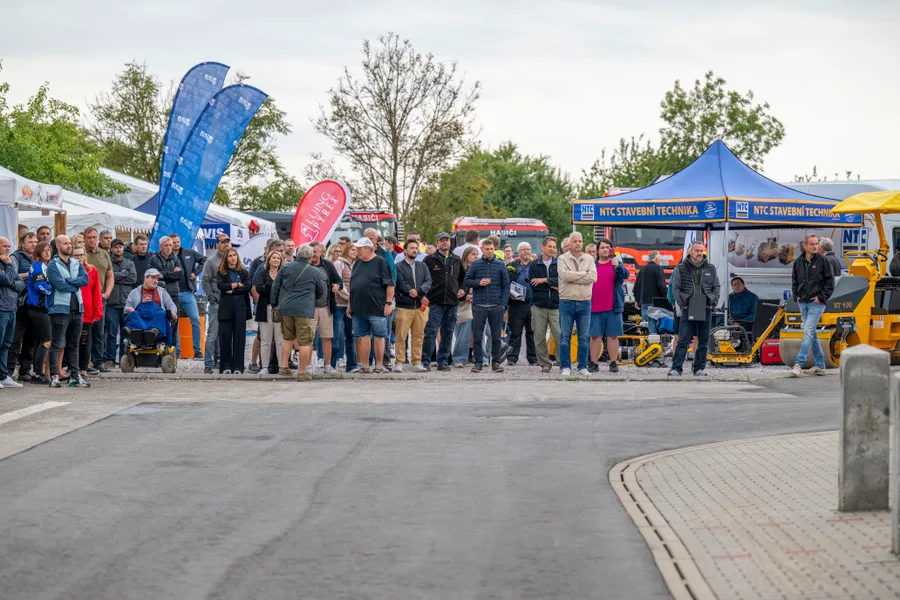 Diváci stojí v řadě podél asfaltové vozovky, za nimi stánky a hasičské vozy