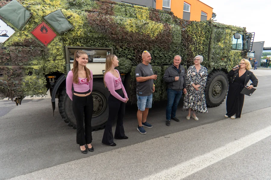 Moderátorka s mikrofonem uvádí skupinu lidí u vozidla pod maskovací sítí