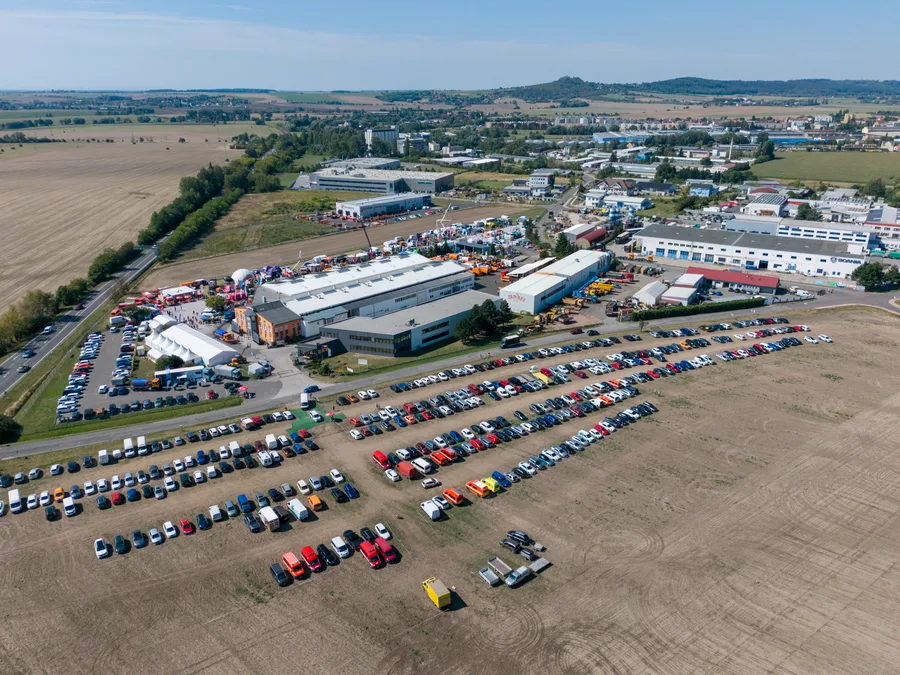 Pole proměněné v parkoviště s řadami aut, za ním haly areálu KOBIT a bílé stany