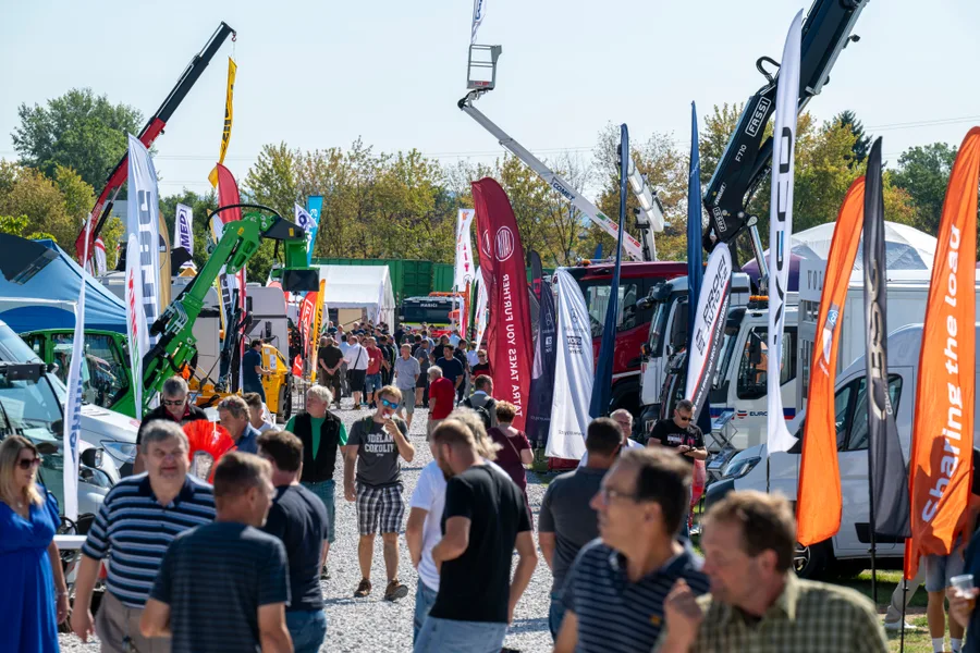 Návštěvníci procházejí uličkou mezi vlajkami, nákladními vozy a vztyčenými hydraulickými rameny